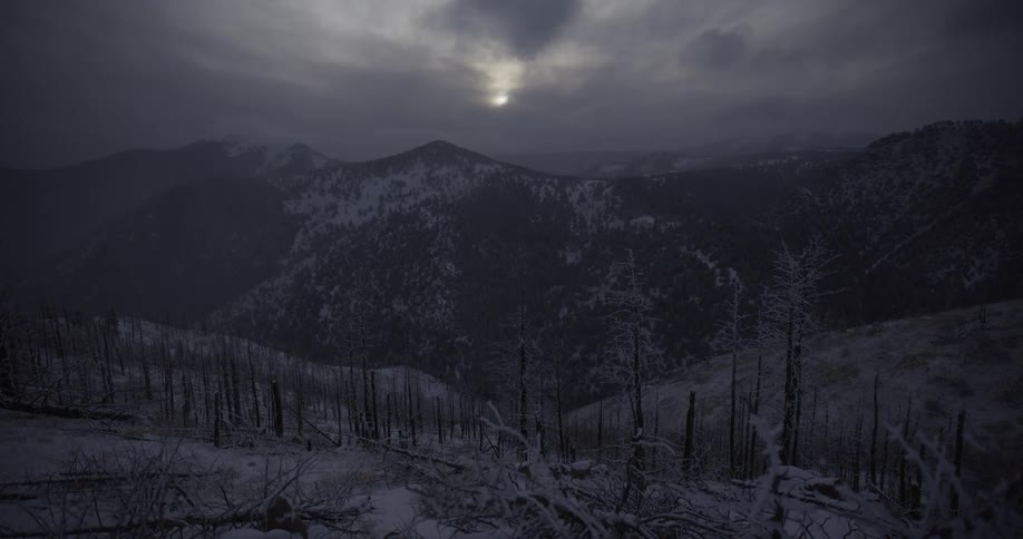 A somber, wide shot of a snow-covered mountain landscape shows the aftermath of a forest fire under a dark, cloudy sky with a faint sun