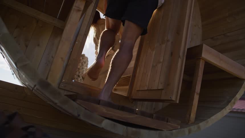 A low-angle slow-motion shot captures the legs and feet of a man and a woman as they enter a wooden barrel sauna and close the door behind them