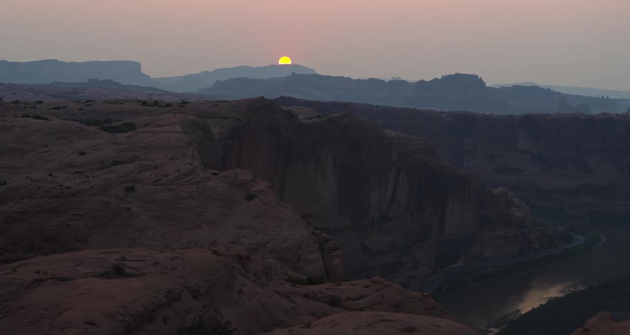 An aerial pullback shot reveals a majestic, hazy sunrise over a deep red rock canyon and a winding river below