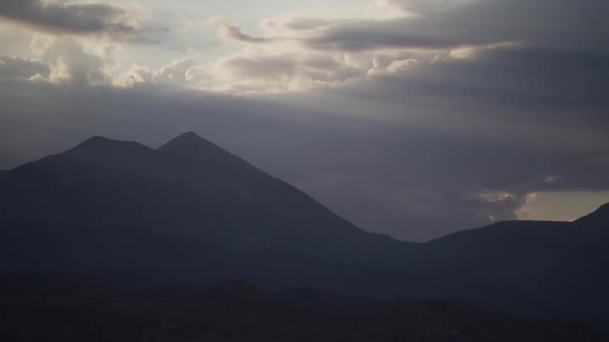 A dramatic timelapse shows clouds moving over a mountain range from dusk to dawn as sunbeams break through and illuminate the landscape