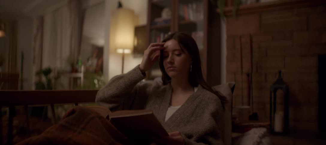 A young woman relaxes on a comfortable couch, wrapped in a plaid blanket while reading a book in her cozy, dimly lit living room