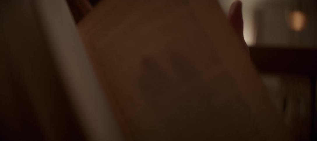 A person wearing a warm sweater turns the page of an old book in a cozy, dimly lit room