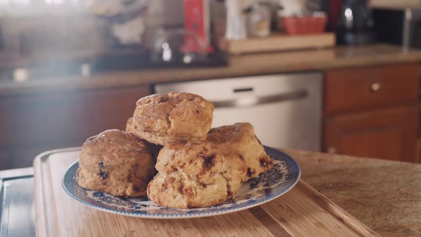A plate of freshly baked scones sits on a wooden cutting board in a sunlit kitchen, with steam gently rising