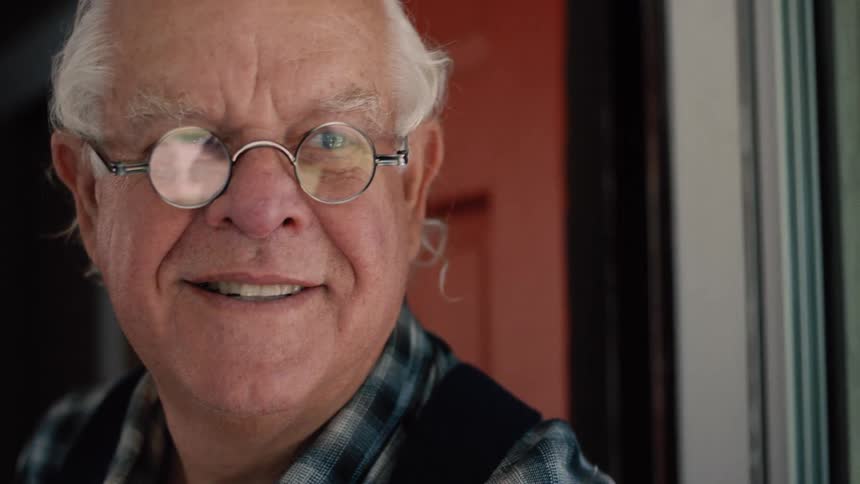 A close-up portrait of a kind elderly man with white hair and round glasses who breaks into a warm, friendly smile