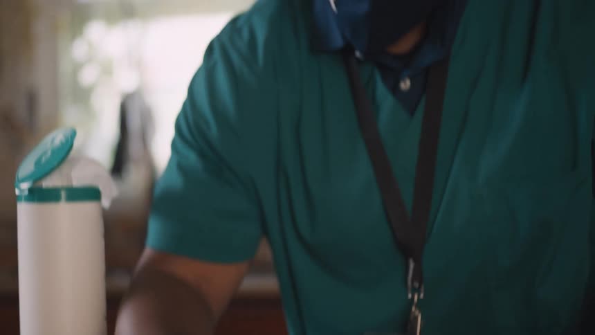 A healthcare worker wearing a face mask and protective gloves carefully sanitizes a countertop with a disinfecting wipe in slow motion