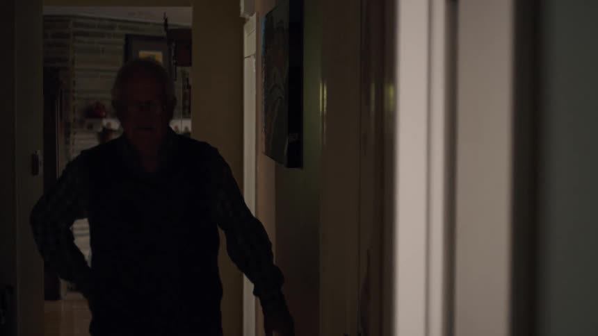 A concerned elderly man walks slowly through a dimly lit hallway at home