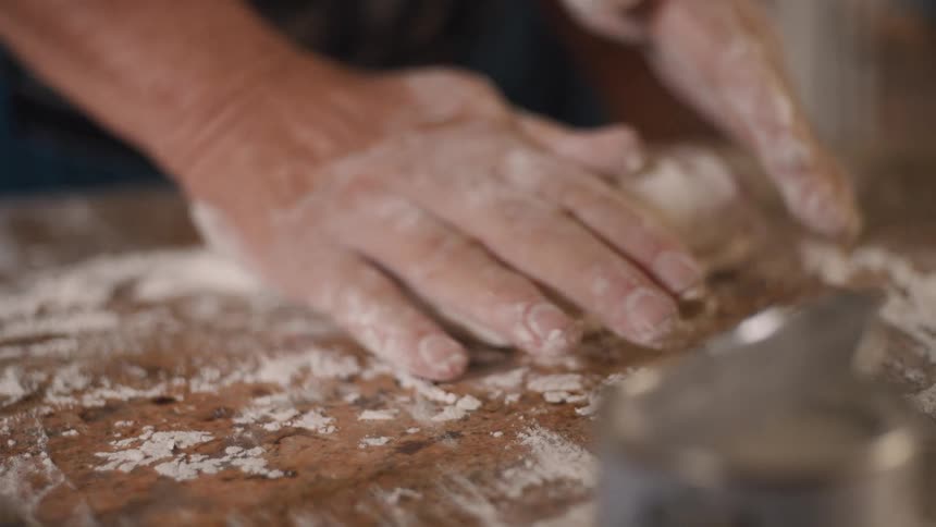 A close-up slow-motion shot shows weathered hands covered in flour expertly kneading soft dough