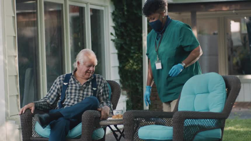 A female caregiver in a mask serves a snack to an elderly man relaxing on a patio, and they share a conversation