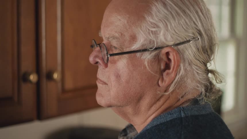 A senior man with long grey hair and glasses pours a fresh cup of coffee into a mug in his cozy kitchen