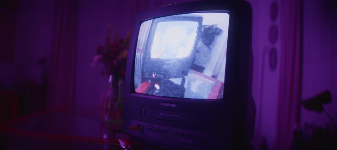 A vintage CRT television with a built-in VCR plays a distorted black and white video as the ambient light in the room changes from blue to purple