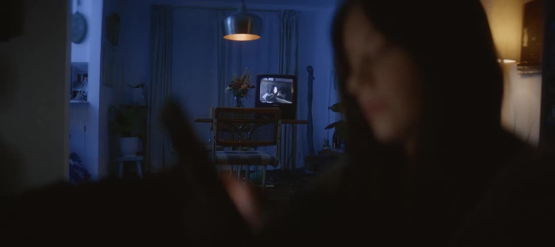 A person scrolls on a device at night, with an old television playing a black and white film in the background of their dimly lit home