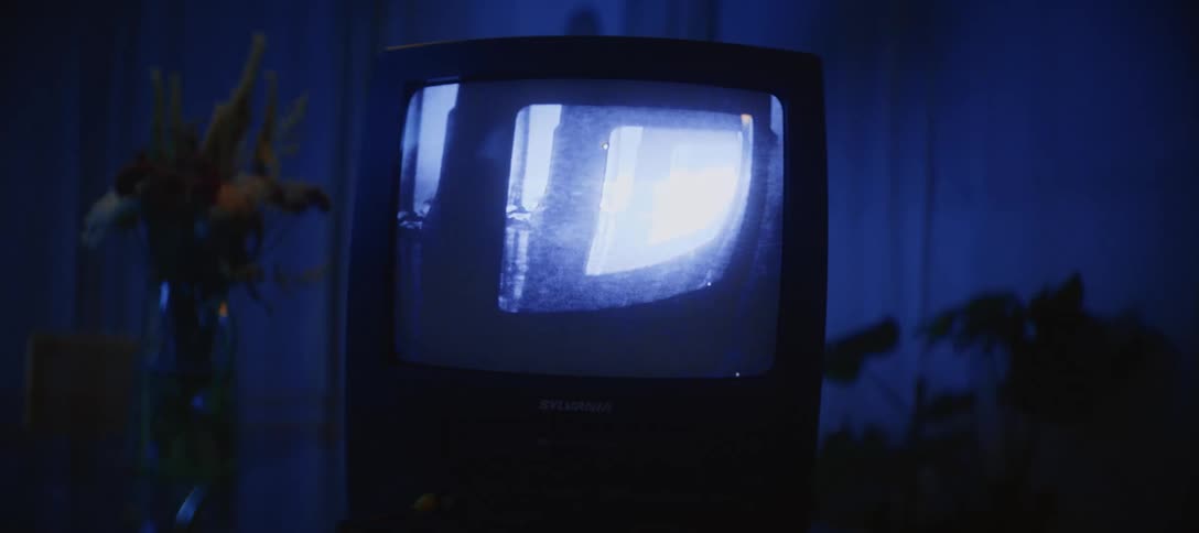 A vintage CRT television and VCR combo glows with a grainy, distorted feedback image in a dark, blue-lit room as the camera slowly pushes in