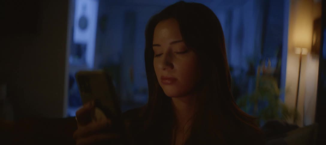 A sad young woman sits alone in a dark room at night, her face illuminated by the glow of her smartphone
