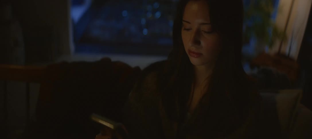 A young woman scrolls on her smartphone in a dark room at night, her face illuminated by the screen's glow