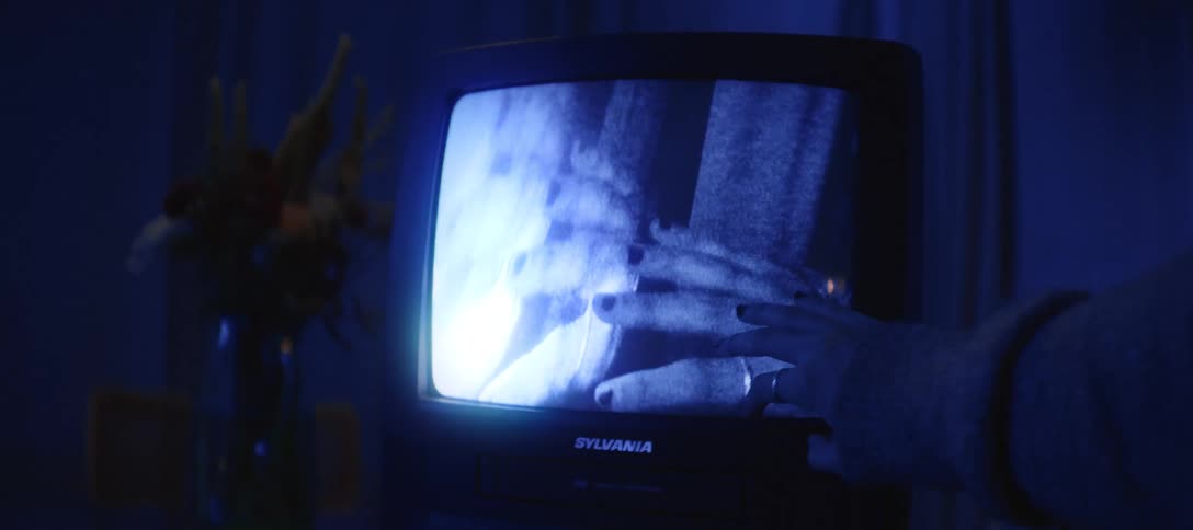 A hand gently touches the screen of an old television displaying a grainy, static image of another hand in a dark, blue-lit room