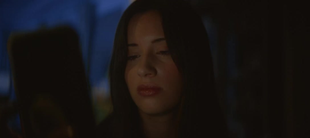 A serious young woman looks down at her phone, her face illuminated by the screen in a dark room at night