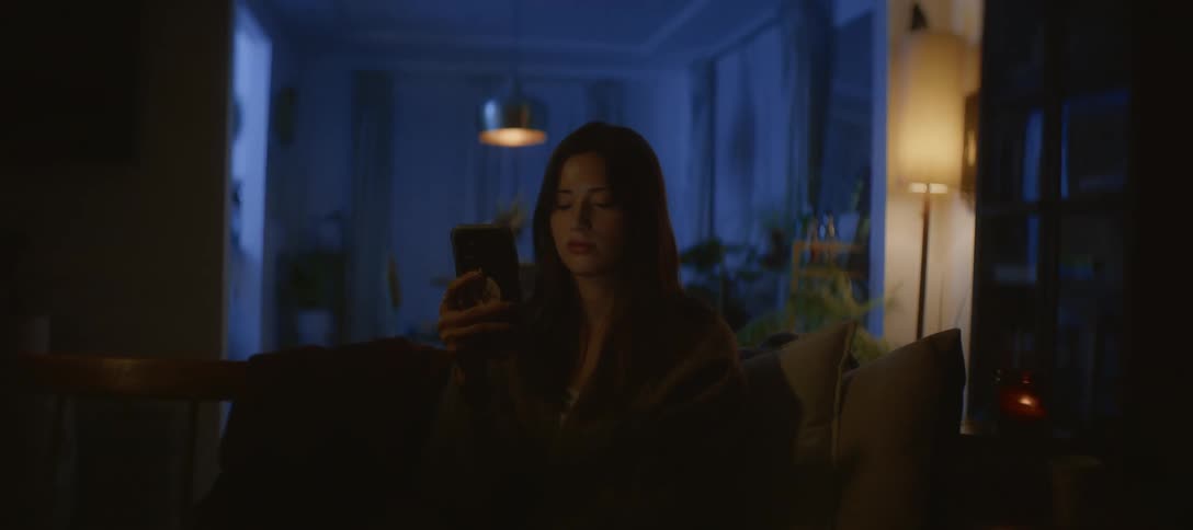 A young woman sits alone on her cozy sofa in a dark room at night, her face illuminated by the glow of her smartphone