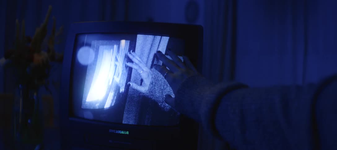 A person's hand reaches out to touch a grainy, surreal image on a vintage CRT television screen in a dark, blue-lit room
