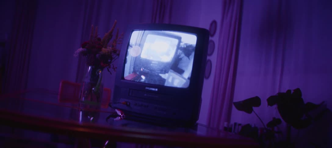 A vintage CRT television set on a table plays a surreal video of itself while the room's lighting cycles through dramatic colors
