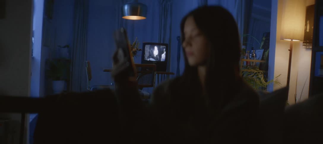 A young woman scrolls through her phone in a dark living room as an old black and white film plays on a vintage television set behind her