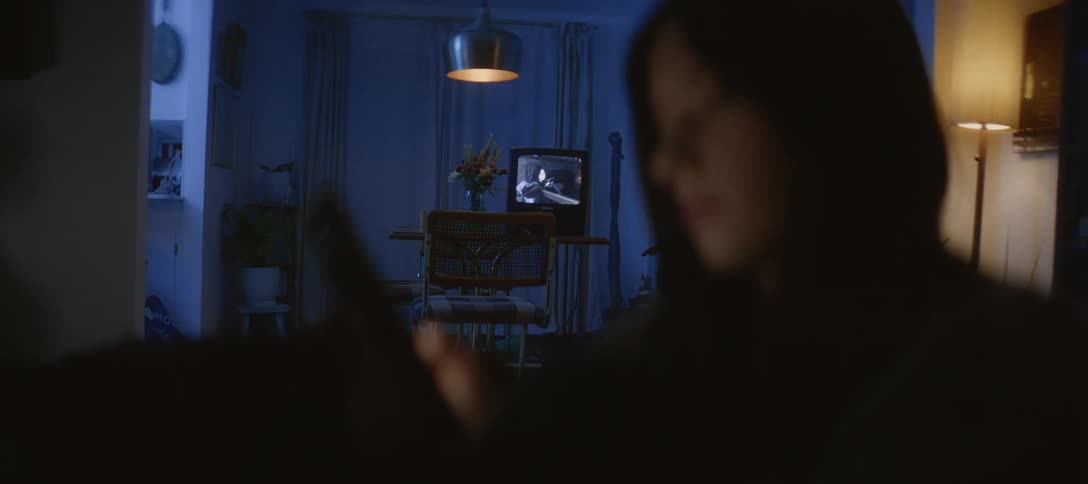 A person sits in a dark room absorbed by their phone while a vintage television plays a black and white film in the background