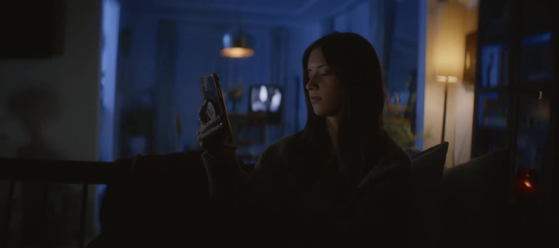A woman's face is illuminated by her phone screen as she sits on a couch in a dark living room at night