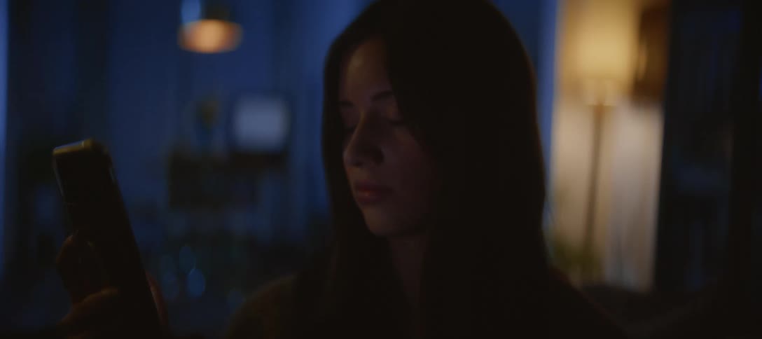 A somber young woman's face is illuminated by her smartphone as she stares at the screen in a dark room at night