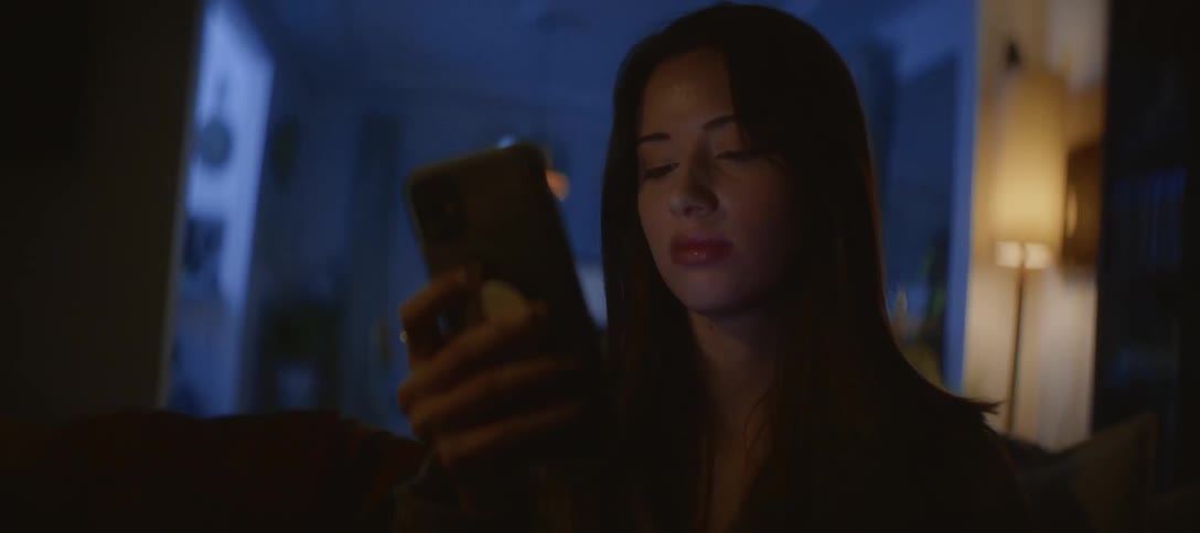 A pensive young woman scrolls through her phone late at night, her face lit by the screen's glow