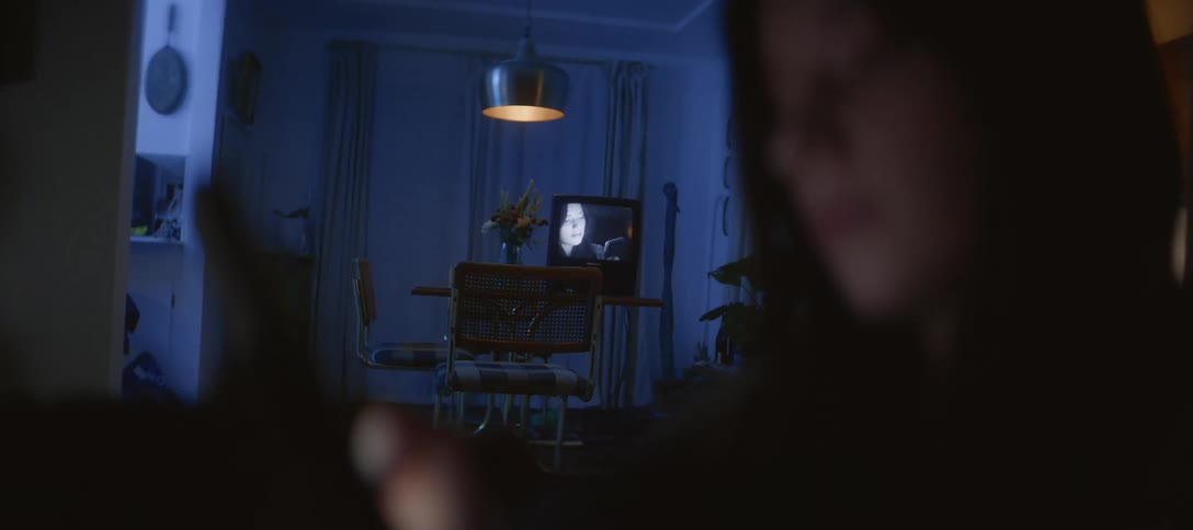 A person sits in a dimly lit room at night, absorbed in their smartphone while a vintage television plays a black and white movie in the background
