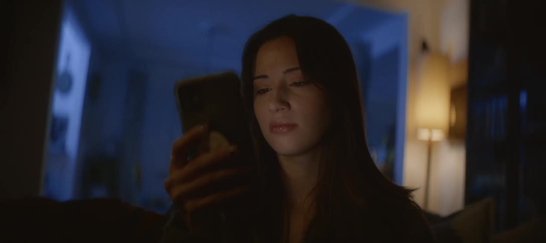 A close-up shot shows a sad young woman looking at her phone in a dark room at night