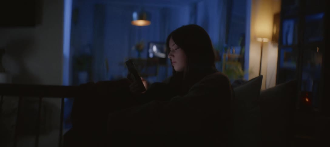 A young woman sits alone in her dark living room at night, the glow from her smartphone screen lighting up her face as she scrolls
