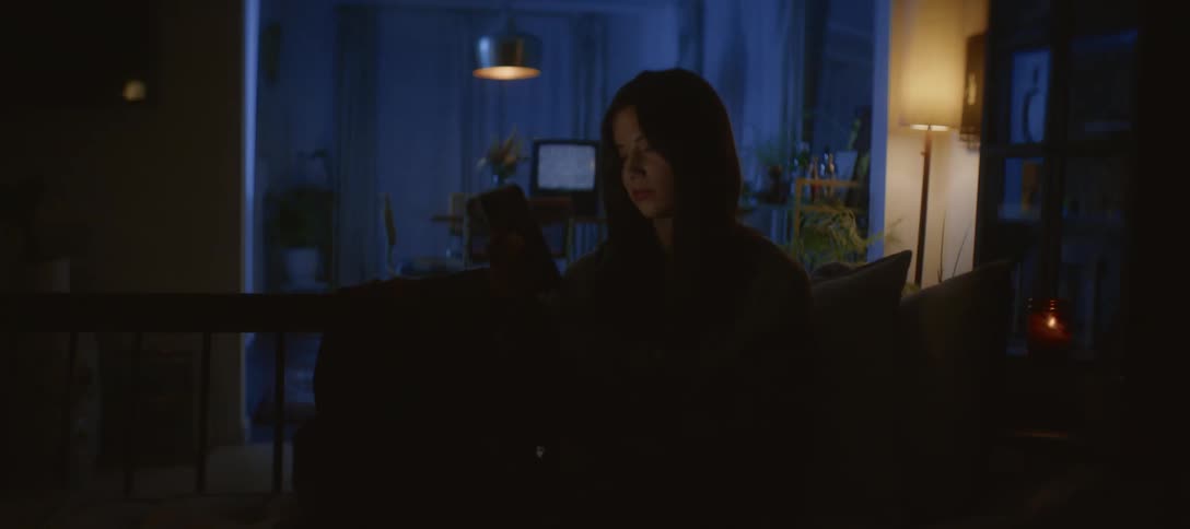 A young woman sits alone on her couch at night, her face lit only by the glow of her smartphone as she scrolls
