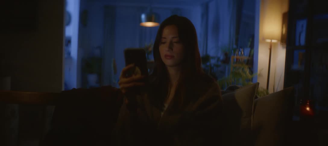 A young woman sits alone on a couch in a dark living room at night, illuminated by the glow of her smartphone screen