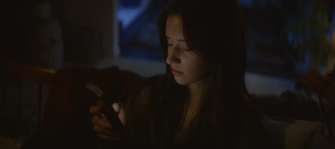 A young woman wrapped in a blanket scrolls on her smartphone in a dark room at night, her face illuminated by the screen's glow