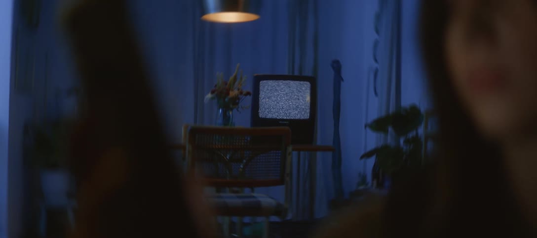 Slow zoom from over a woman's shoulder onto a vintage television with static in a dark, blue-lit room