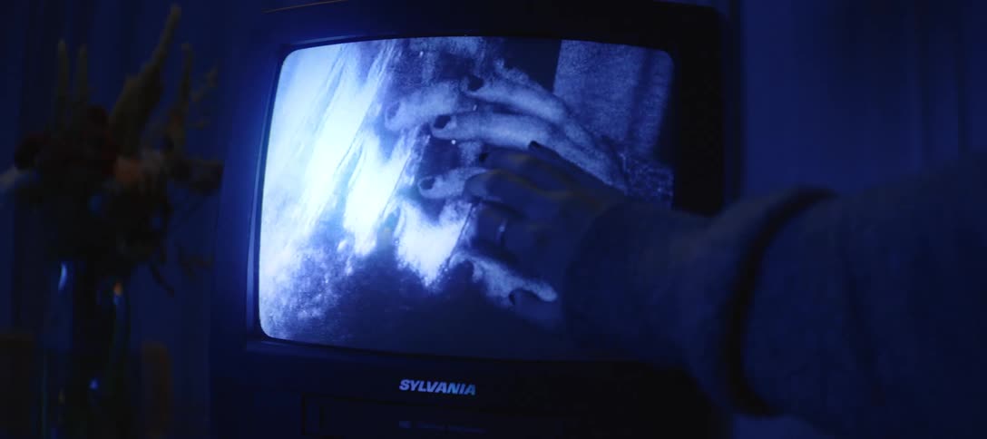 A hand gently touches the screen of an old television displaying a ghostly, static-filled image of another hand in a dark, blue-lit room