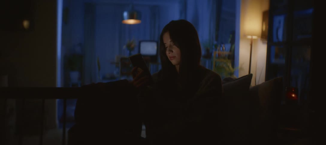 A young woman wearing a hoodie scrolls on her smartphone, her face illuminated by the screen's glow in a dark living room at night