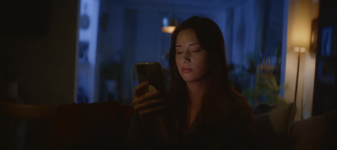 A woman scrolls on her smartphone in a dark, cozy living room at night, her face illuminated by the screen's glow