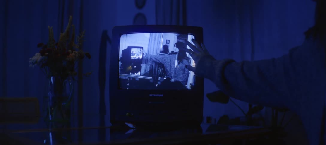 A hand gently touches the screen of an old television playing a black and white home video in a dark, blue-lit room