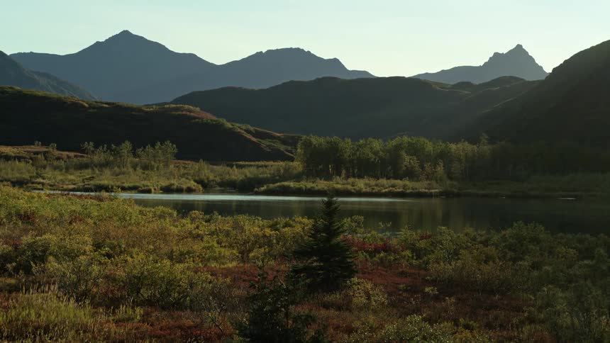 A slow push-in reveals a calm lake in a remote valley surrounded by silhouetted mountains and lush vegetation at sunrise