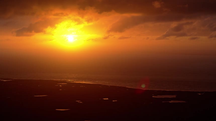 A brilliant golden sun sets over the ocean, casting an orange glow across the sky and reflecting on the silhouetted coastline