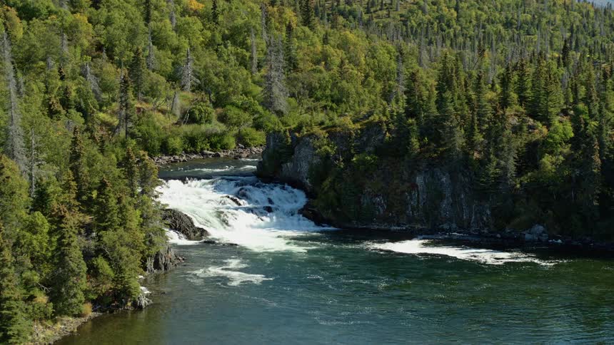 An aerial view slowly approaches a powerful waterfall cascading down a river in a dense, remote forest wilderness