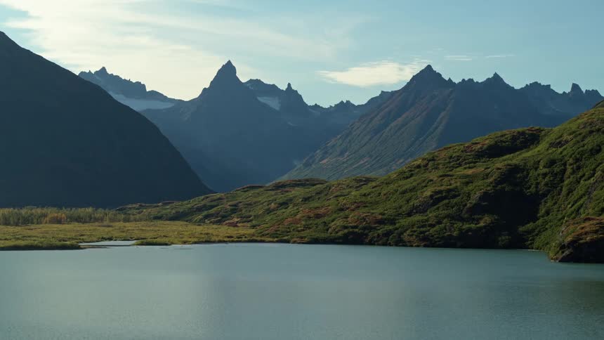 An aerial view slowly moves over a pristine lake towards a lush green valley and a majestic snow-capped mountain range
