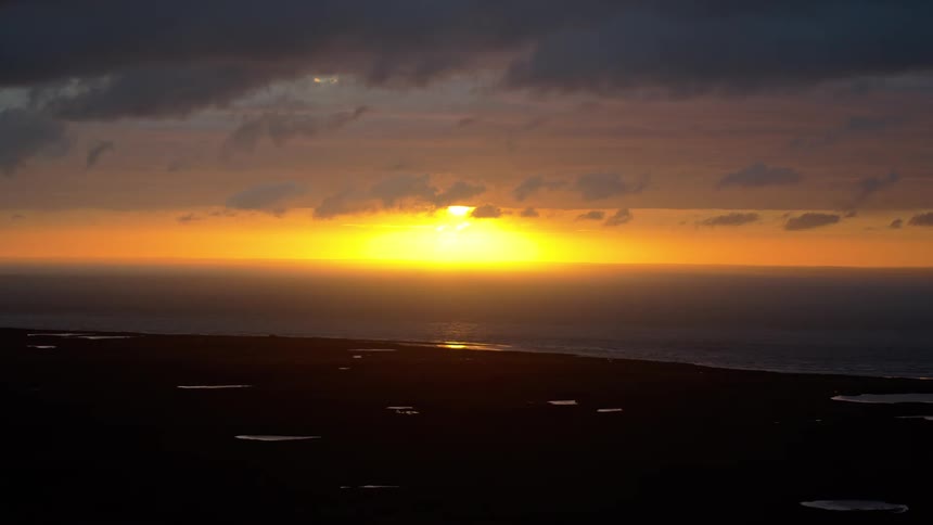 A brilliant golden sunset pierces through dark clouds over the ocean, with a silhouetted landscape in the foreground