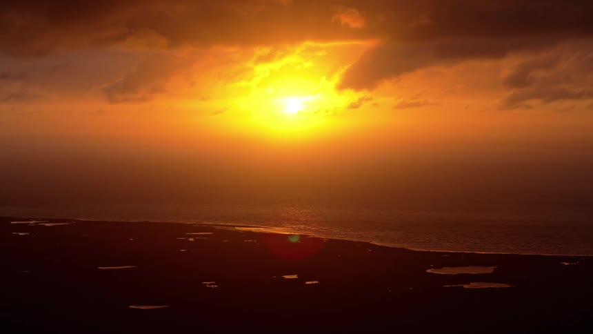 A dramatic orange sunset glows brightly through clouds over a silhouetted coastline and the ocean