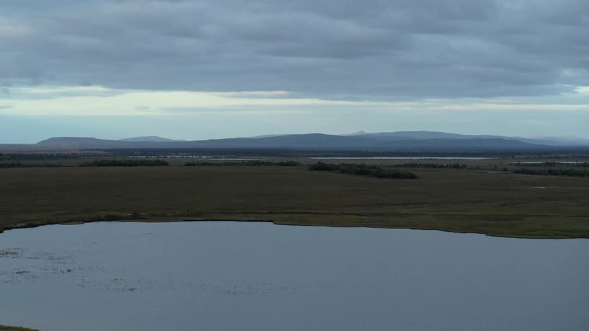 Aerial view rises over a tranquil lake in a vast tundra landscape with distant mountains under a cloudy sky