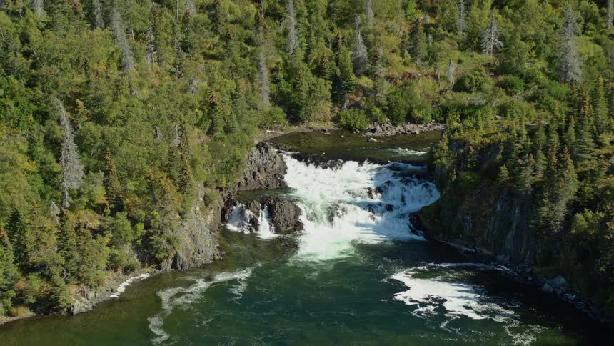 An aerial view pulls back from a powerful waterfall cascading over rocks in a remote river canyon surrounded by a dense forest