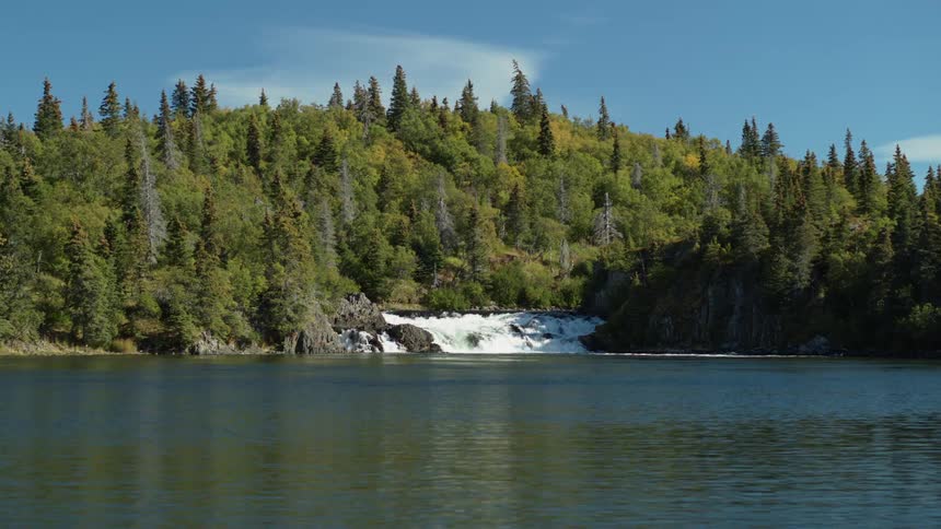 A beautiful waterfall cascades into a tranquil river surrounded by a lush green forest on a sunny day