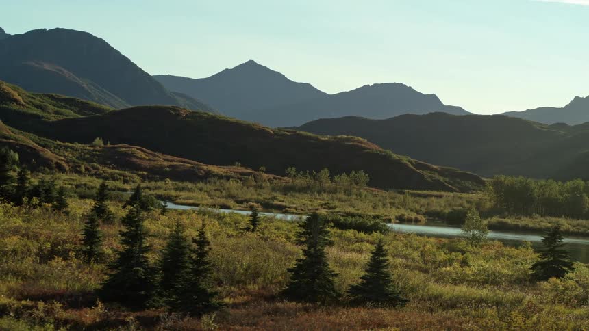 A slow tracking shot reveals a serene river winding through a vast, sun-kissed valley at the base of majestic rolling mountains