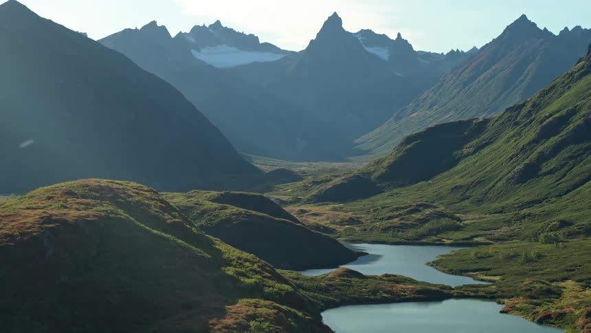 Sunlight illuminates a stunning, remote mountain valley with a tranquil lake and rugged, snow-capped peaks in the distance